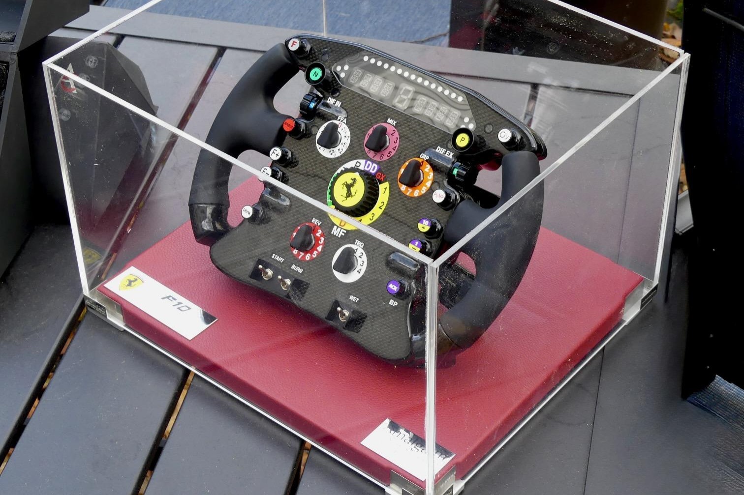 This replica Ferrari F10 steering wheel was produced by Amalgam Collection of Bristol, England, in 2010 under license by Ferrari, and it is displayed in a clear plastic box with a red base. The wheel is fitted with a faux screen, shift paddles, LED lights, sw…